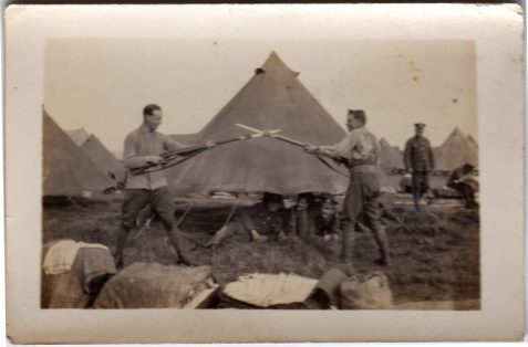 Photograph of two soldiers with bayonets crossed and pointed at each other outside tent watched by several others