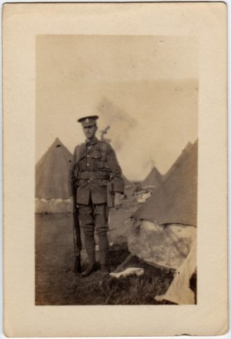 Photograph showing a soldier standing with his rifle outside tents