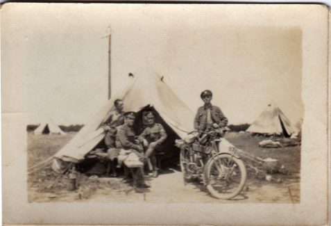 Photograph of five uniformed men outside a tent, one standing next to a motorbike, other tents are visible in the background