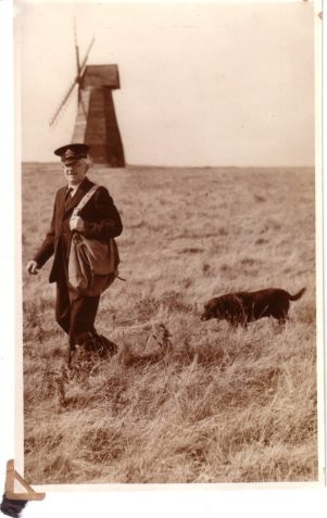 Photograph of John Leech and dog in front of Rottingdean windmill in a postman's uniform carrying a postbag