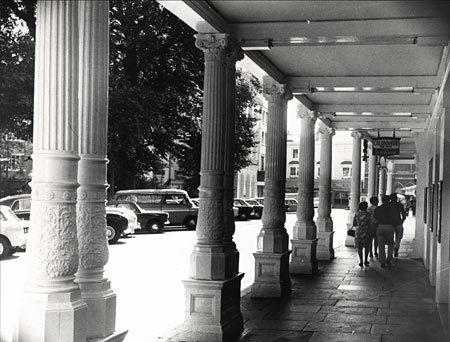 This photographic print was made by the Borough Surveyor's department. It is a view of New Road, Brighton, taken from within the Theatre Royal Colonnade on the west side of the street.  This colonnade was originally constructed in 1806-7. The section featured in the photograph was replaced in 1894 when the Theatre Royal was reconstructed. | Reproduced courtesy of Royal Pavilion, Libraries & Museums, Brighton & Hove
