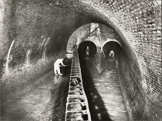 This is a photographic print of the Brighton & Hove intercepting sewer. It shows the meeting point of the London Road and Lewes Road Valley sewers. Two workmen can be seen standing in the tunnel entrances at the rear of the sewer. Water is running down through these tunnels while another workman attends to the overflow area on the left of the photograph.  The tunnels visible at the rear of the photograph are approximately 2.5 metres high. If the water was too high, it would fill into the 27 metre overflow channel. The overflow water would eventually emerge at the storm water outfall by the Albion Groyne near the Palace Pier. The channel itself is protected by the metal scum board visible in the centre of the photograph.  This photograph was commissioned by the Borough Surveyor's department of Brighton Borough Council. It was probably taken to record improvements made to the structure. | Reproduced courtesy of Royal Pavilion & Museums, Brighton & Hove