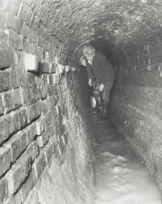 This is a photographic print of Marine Parade Sewer, Brighton. It is a view of the inside of the narrow tunnel, taken at a point east of the junction with the Old Steine. Two workmen can be seen inspecting the brickwork.  This photograph was commissioned by the Borough Surveyor's department. A note accompanying the photograph states that the 'defective brickwork was replaced, the whole repointed and rendered to the shoulders'. | Reproduced courtesy of Royal Pavilion & Museums, Brighton & Hove