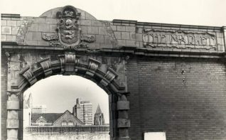Facade of the Market Buildings, c. 1970s: An arched gateway which had led to market buildings built in 1900-1901 by the borough engineer Francis May in red brick and terracotta design in three sections. Each building had an iron roof and arched glass, selling fruit, vegetables and flowers. They closed in 1938 when the Circus Street Municipal Market opened, and was demolished. This archway and Market Street disappeared in the development of Bartholomew Square which opened in 1987 | Image reproduced with kind permission from Brighton and Hove in Pictures by Brighton and Hove City Council